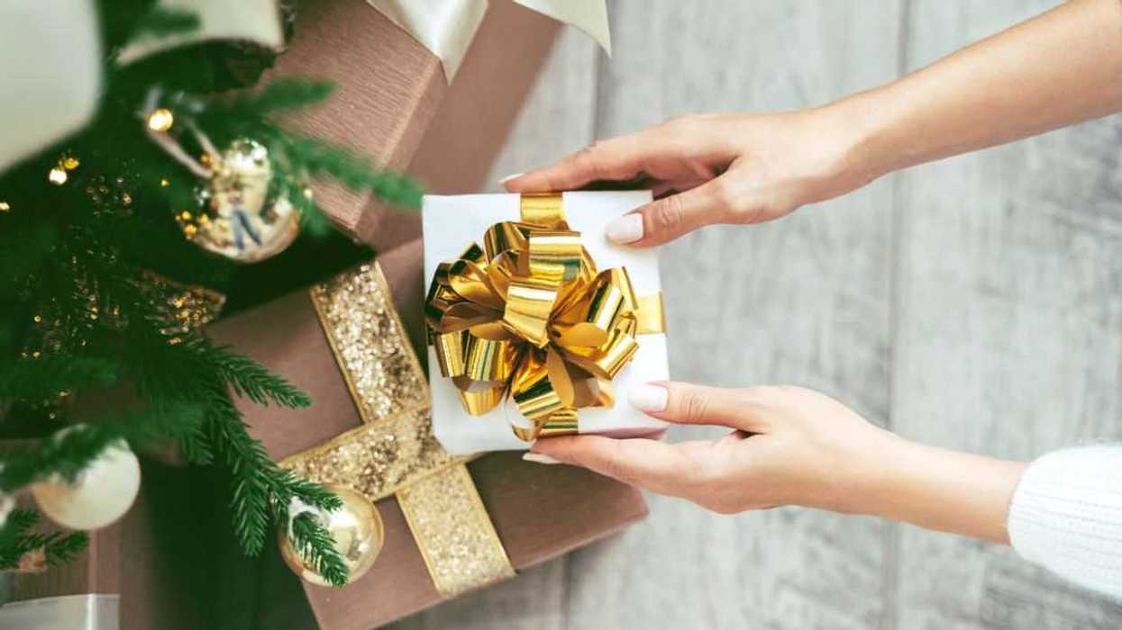 Tender woman hands putting white wrapped gift box with golden bow tie under the Christmas tree to other presents.