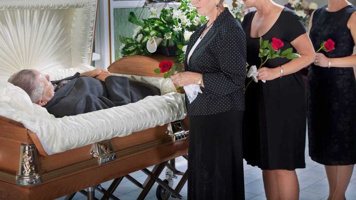 Three women standing by a man's body laying in a coffin.
