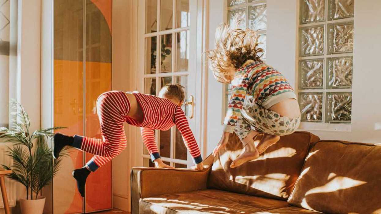 Two cute kids wearing pyjamas bounce on a brown leather couch in a sunny domestic room.