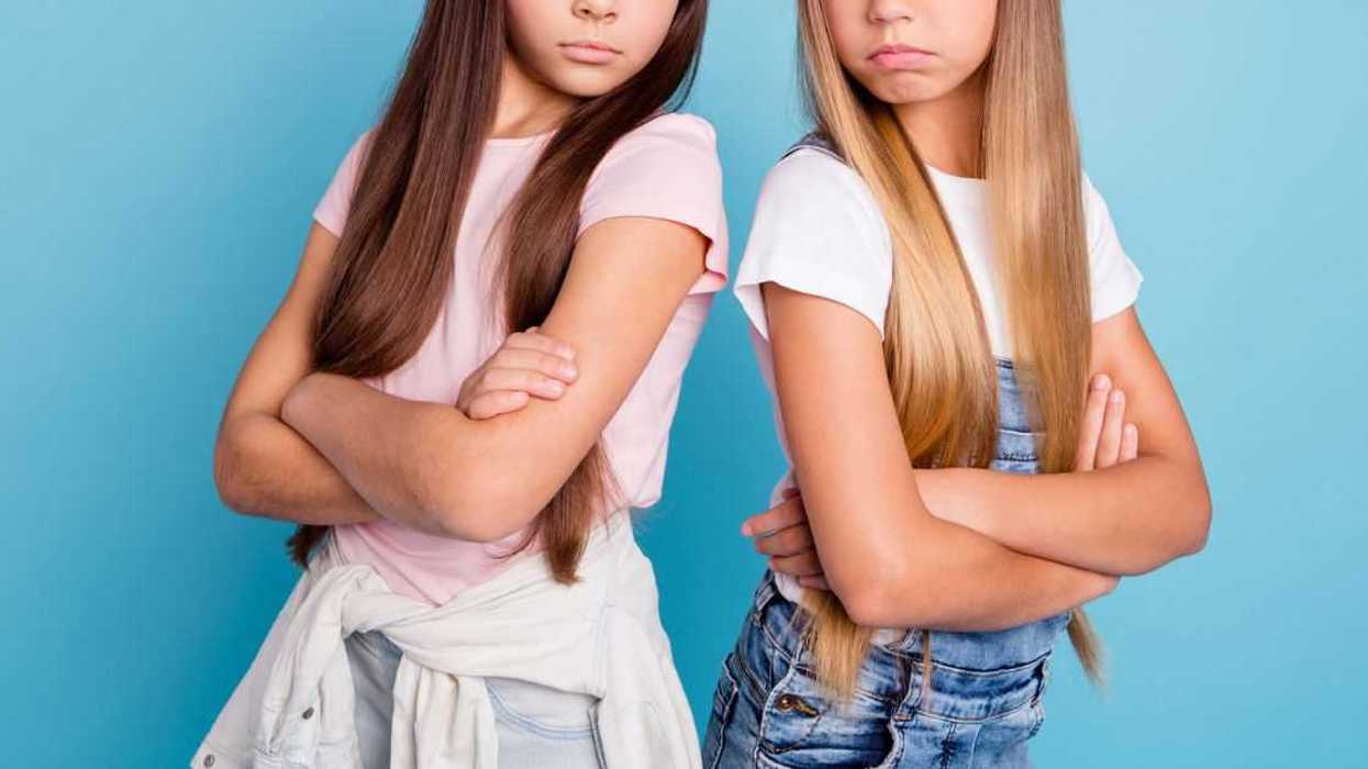 Two frowning adolescent girls stand back to back with their arms folded.