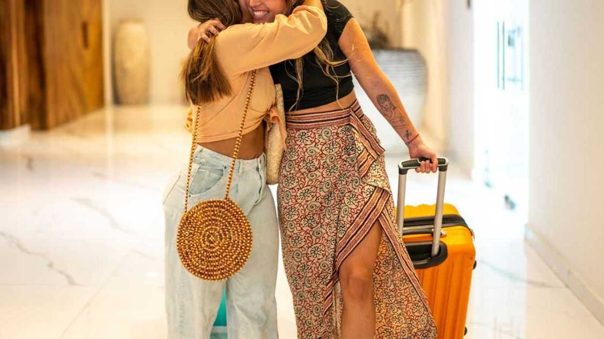Two young female friends embrace, one holds a suitcase