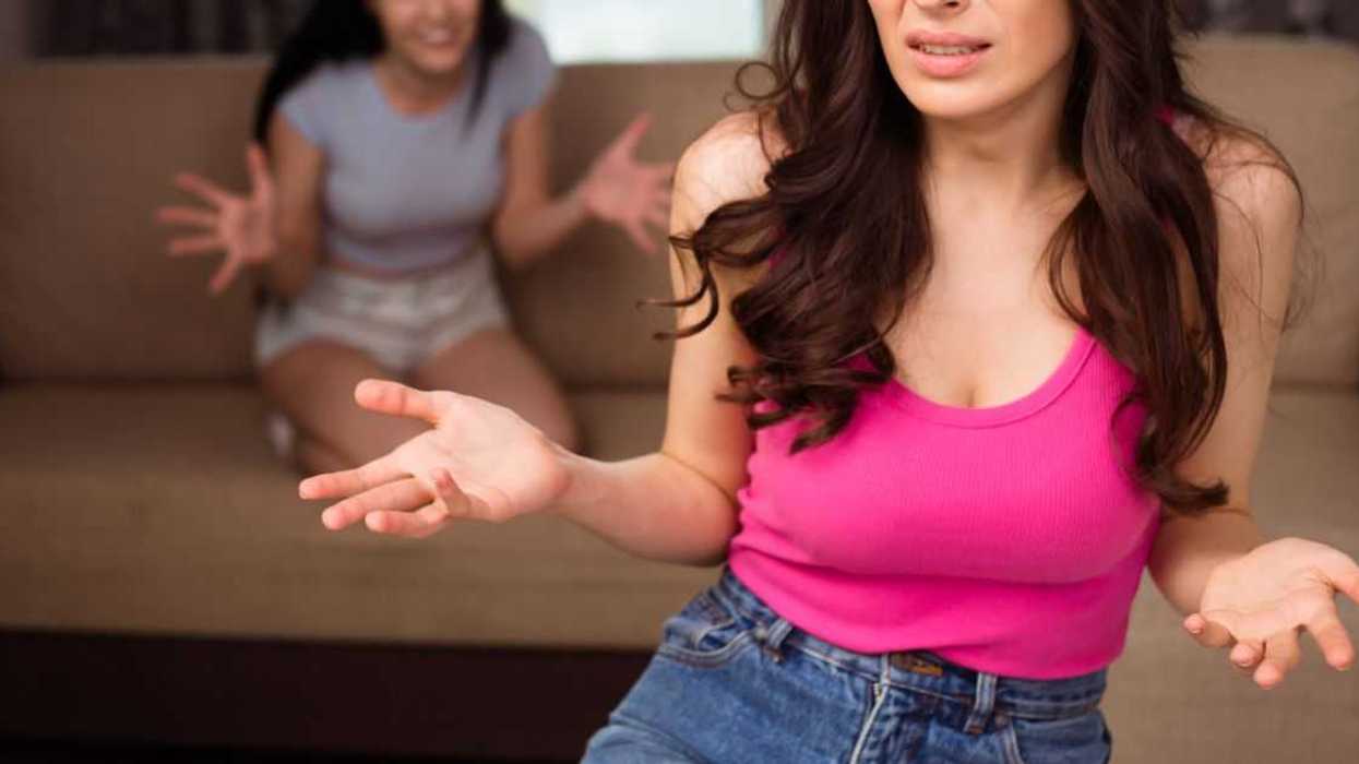 Two young girlfriends in casual wear quarrel and shout at each other in the apartment.