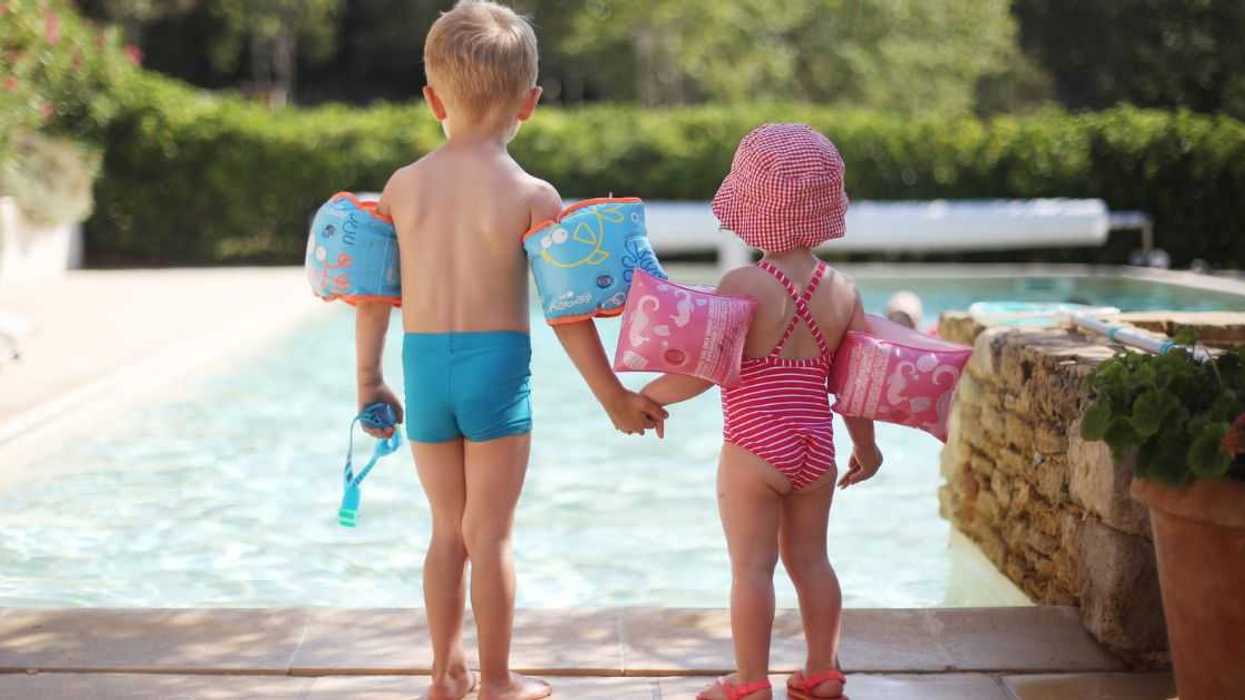 Two young kids, a boy and girl, stand at a pool wearing arm floaties