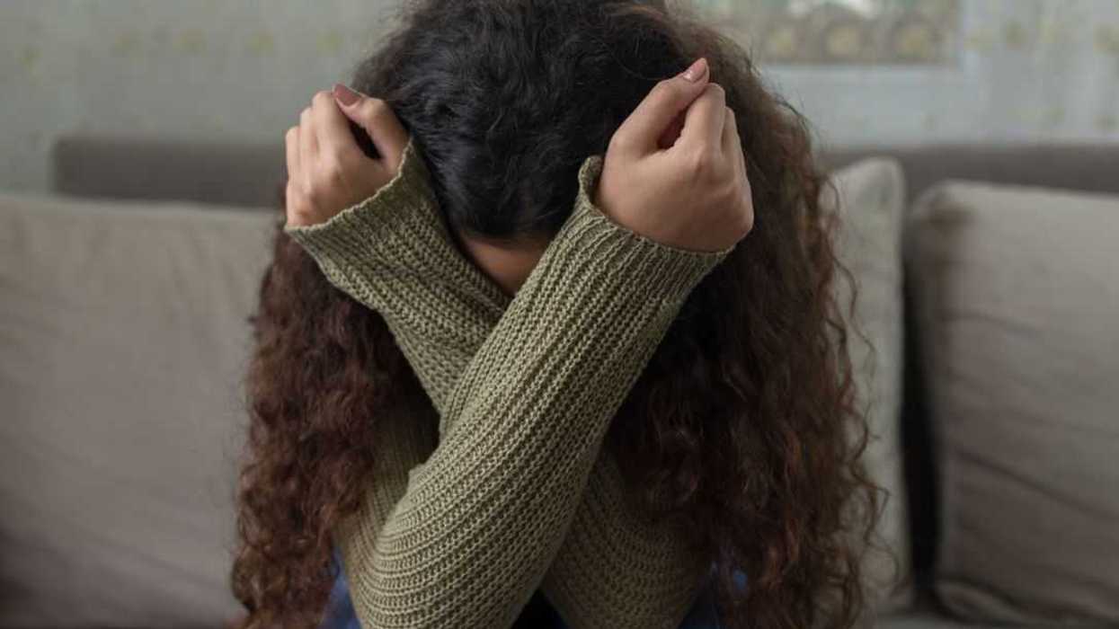Unhappy girl on sofa sits with her arms crossed over head. Her face is hidden.