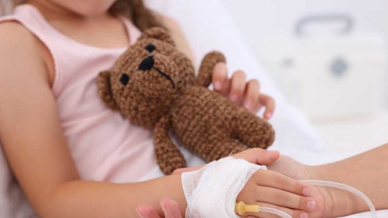Unidentifiable mother and daughter hold hands as daughter is in a hospital bed, hooked up to an IV drip. Close-up of the little girl's light brown teddy bear.