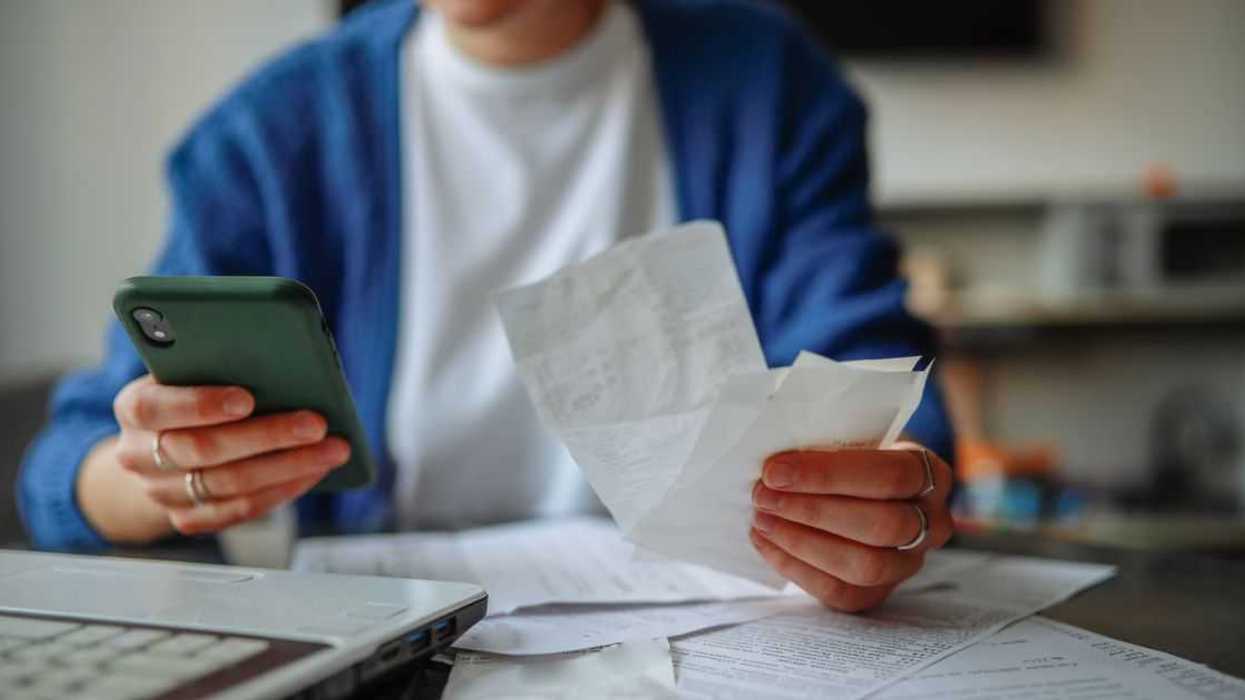 Unrecognizable woman with a smartphone, laptop, and checks at a table at home.