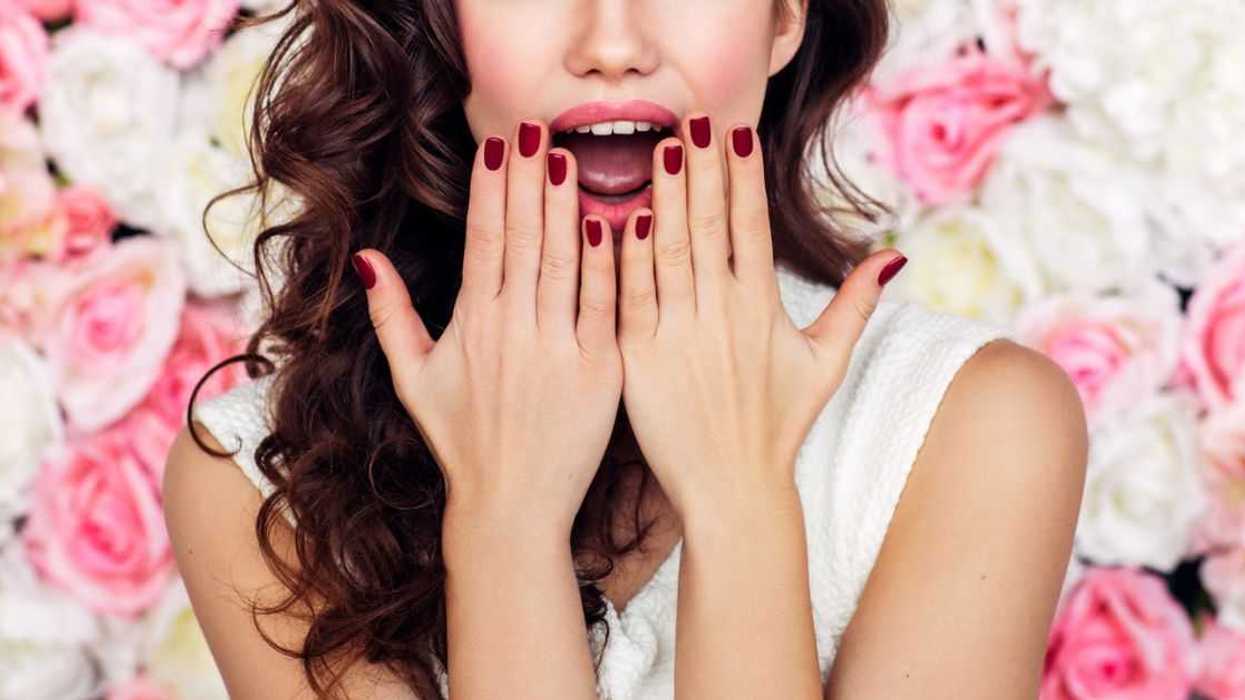While standing against a wall of flowers a bride is shocked with her hands pressing against her cheeks
