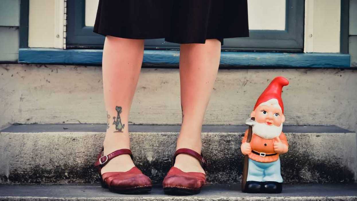 Woman wearing red shoes, tattoos standing next to a garden gnome.