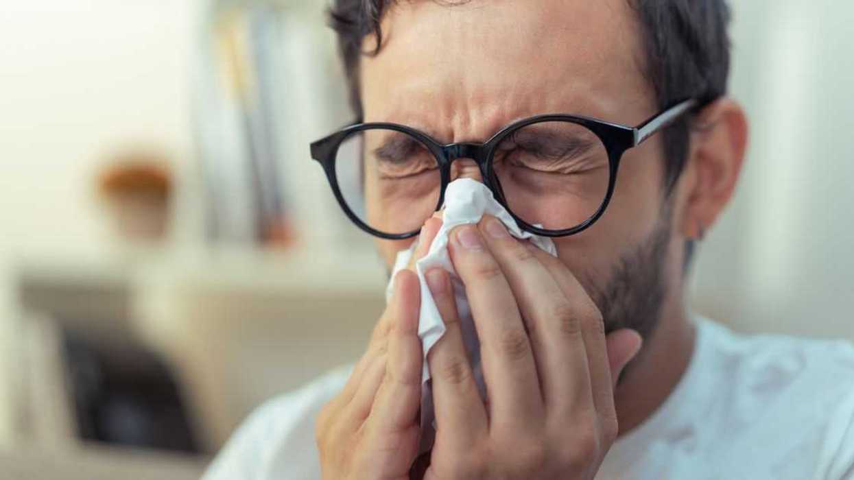 Young man with glasses sneezing, wiping his nose with a piece of tissue paper.