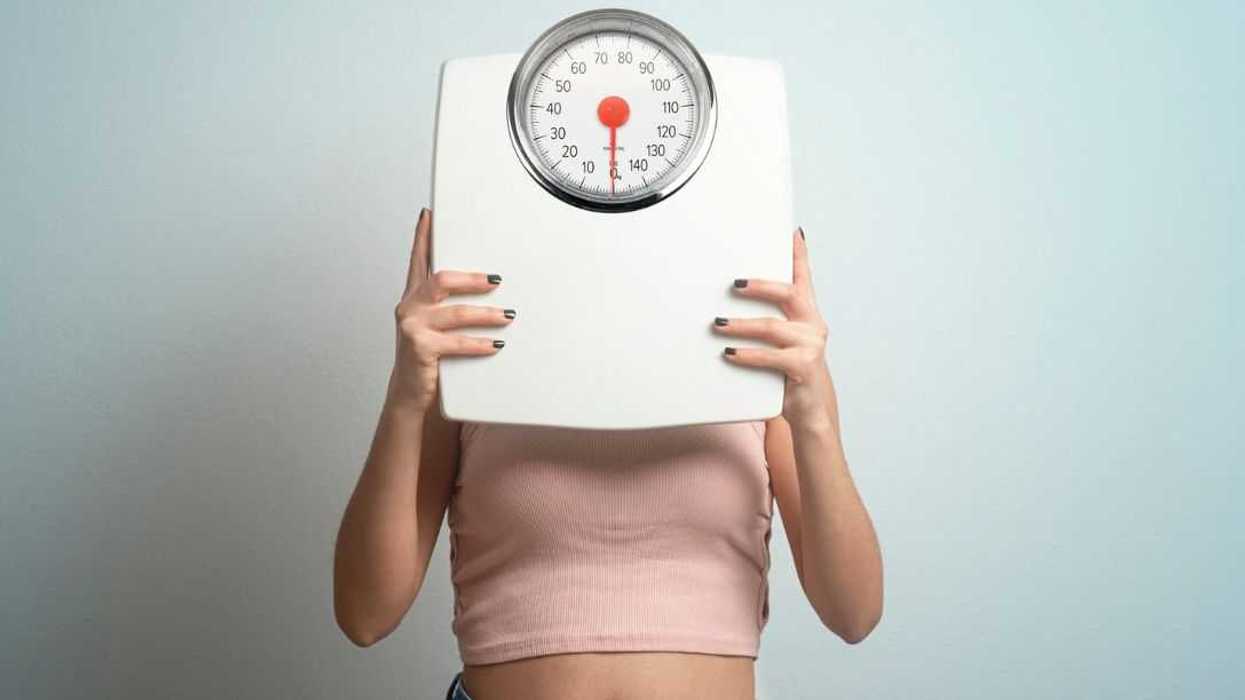 Young woman hides behind bathroom scales. Studio shot.