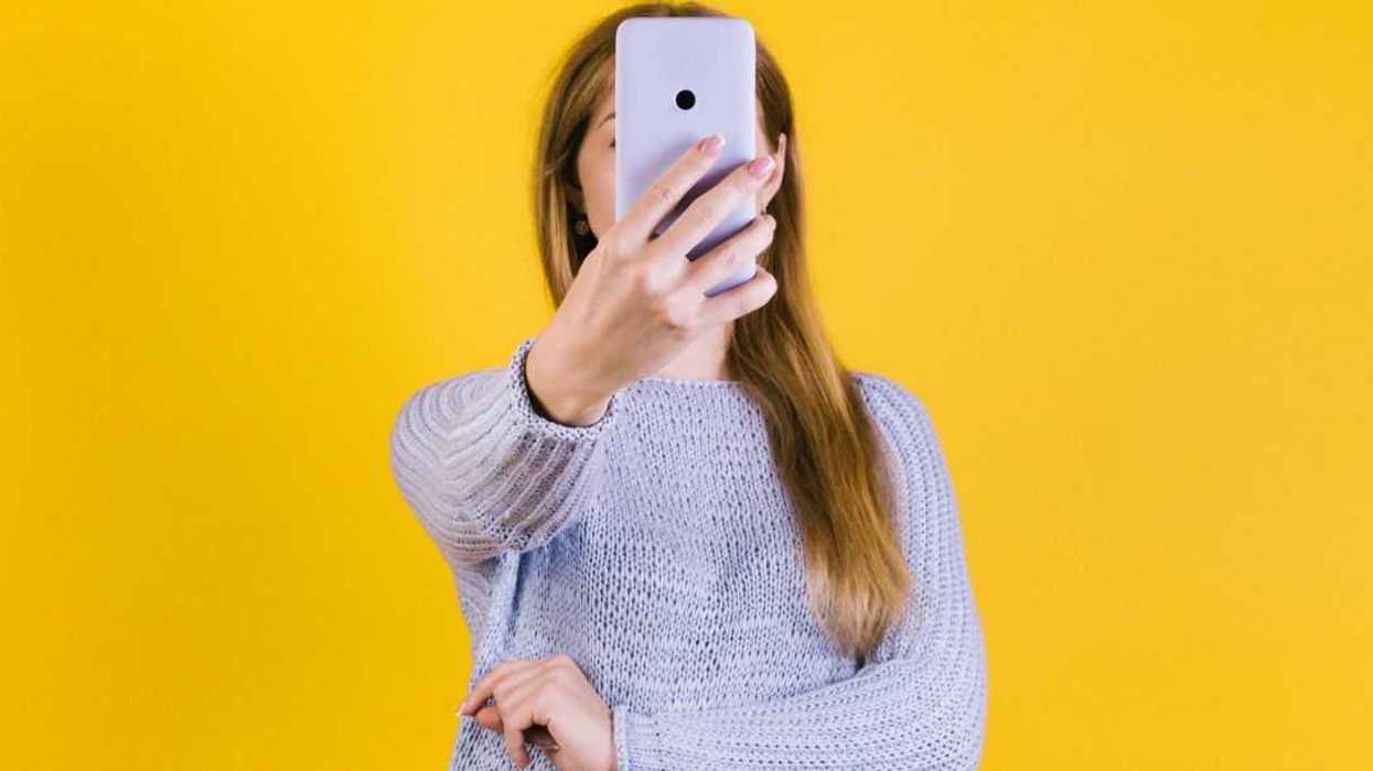 Young woman holding smartphone in front of her face against a yellow backdrop.