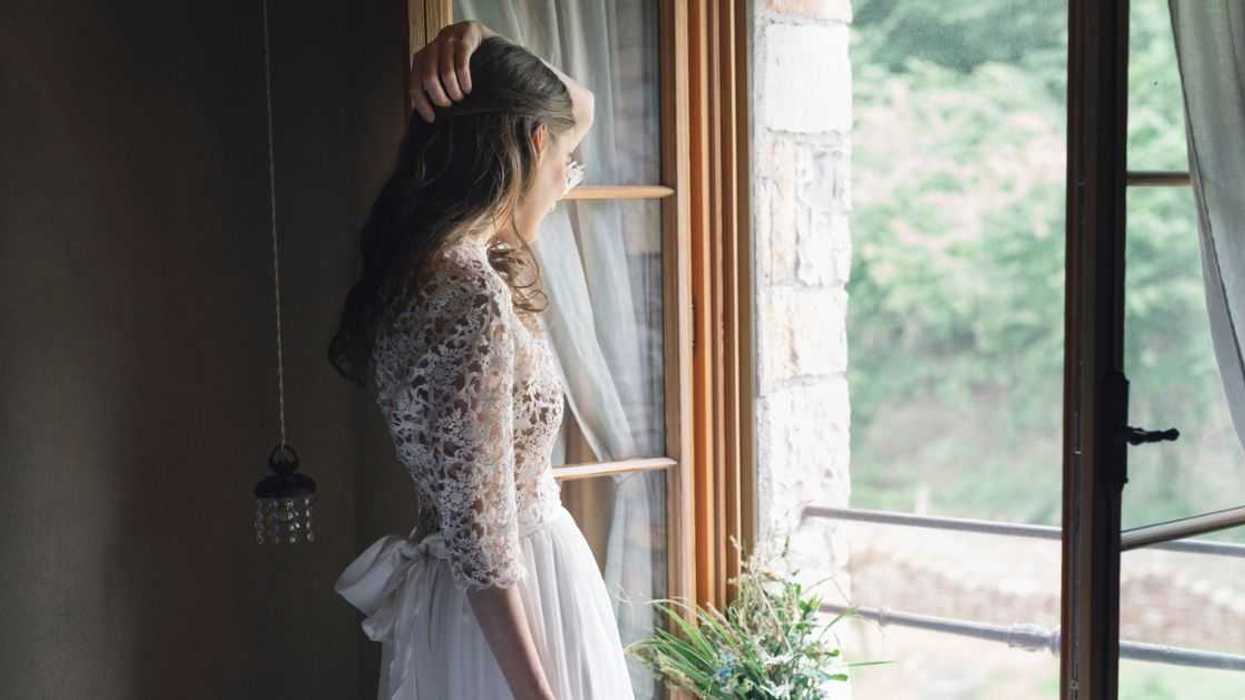Young woman in elegant wedding dress holding bouquet looking out of the window.