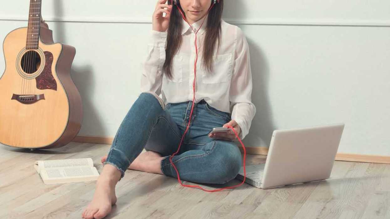 Young woman sitting on wooden floor listening to music through headphones. Next to her are a laptop, a smartphone and a guitar.
