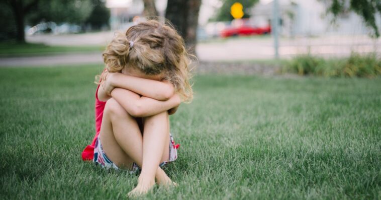 A young girl sits in the grass with her face collapsed into her arms