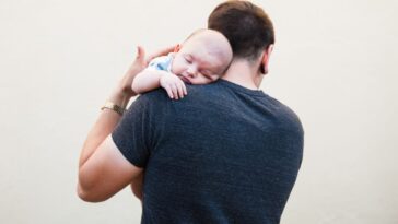 A man with his back to us caresses a baby's back while it rests on his shoulder