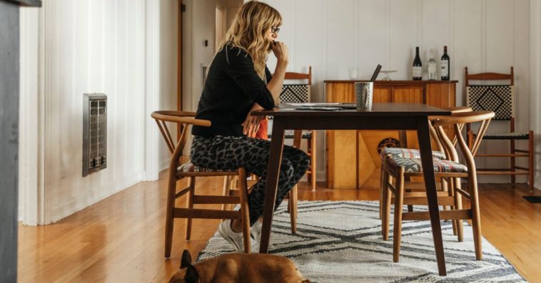 A woman sits alone at her dining table with her dog at her feet