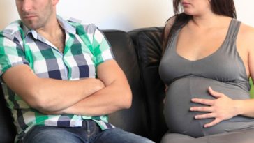 A married couple, the woman pregnant, sit on a couch angry