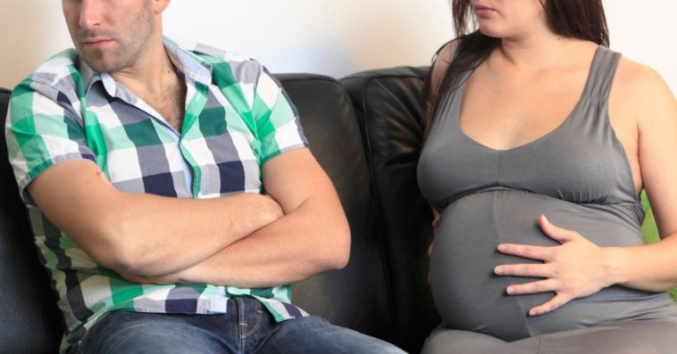 A married couple, the woman pregnant, sit on a couch angry
