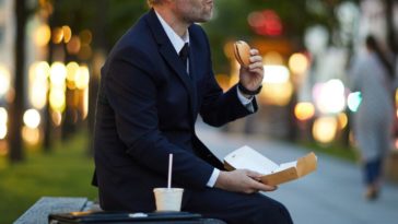 A guy in formal attire sits at a bench, eating fast food.