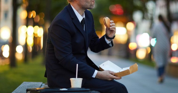 A guy in formal attire sits at a bench, eating fast food.