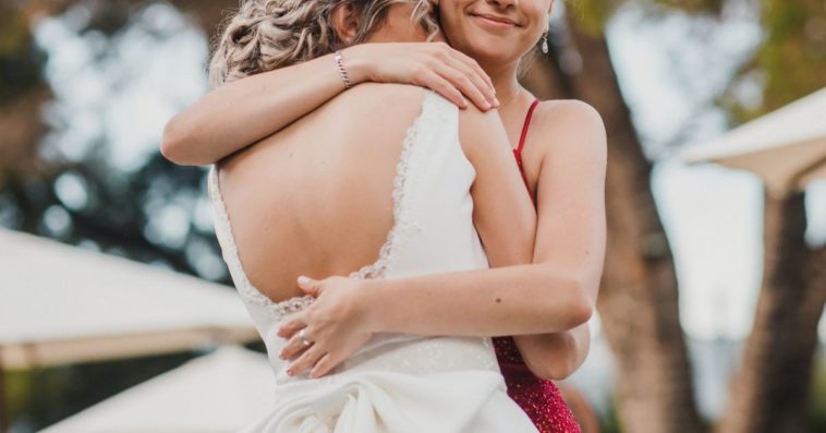 A bridesmaid in a red dress hugs a bride