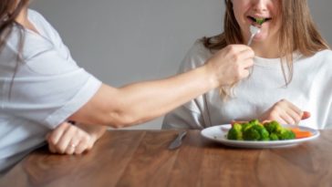 A woman force feeds a crying young woman broccoli