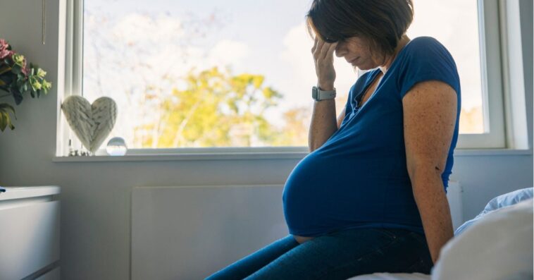 A pregnant woman sits on the edge of a bed seemingly in despair