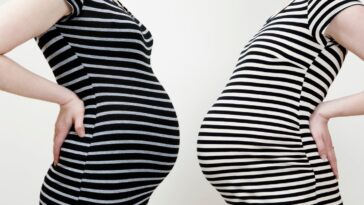Two pregnant women stand stomach to stomach in similar dresses