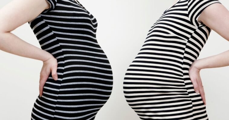 Two pregnant women stand stomach to stomach in similar dresses
