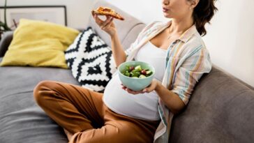 A pregnant woman eats pizza and salad on a couch
