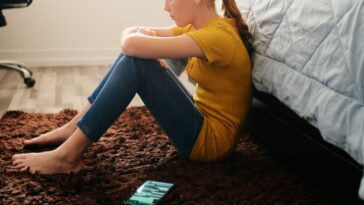 A teen girl sits on a bedroom floor, sulking