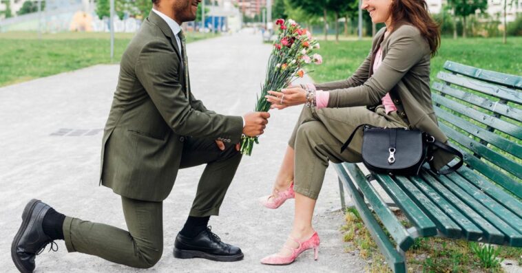 A man in a green suit gives flowers and proposes to a woman on a park bench