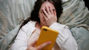 A woman lays in bed, looks at her phone, her hand covers her face