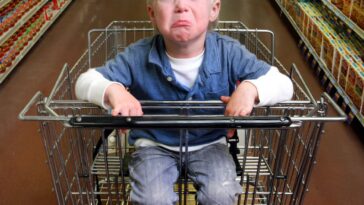 A very upset young boy sits in a wagon in a grocery aisle