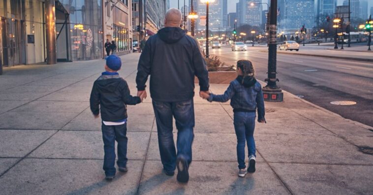 A man walks down the street holding the hands of two children