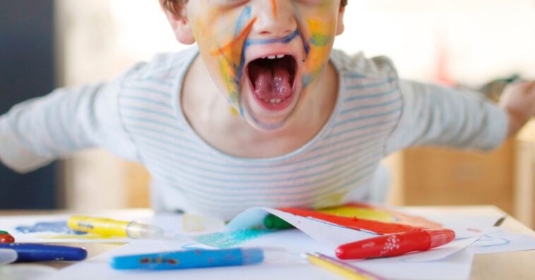 A young child with his face covered in colorful paints yells into the camera