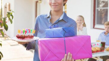 A teen boy holds a birthday cake and a few presents at a party