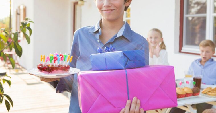 A teen boy holds a birthday cake and a few presents at a party