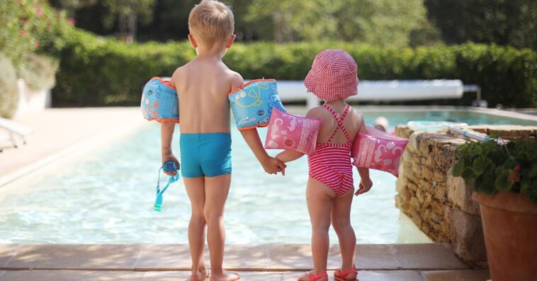 Two young kids, a boy and girl, stand at a pool wearing arm floaties