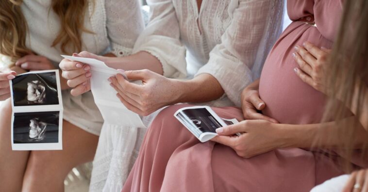 A very pregnant woman is surrounded by women at a baby shower as they look at sonograms