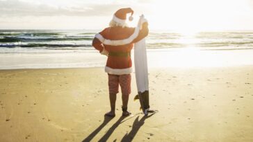A Santa stands on a beach on a sunny day, holding a surfboard