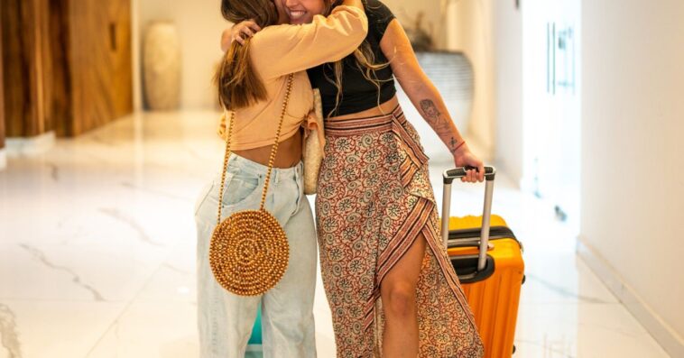 Two young female friends embrace, one holds a suitcase