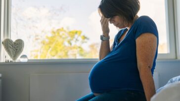 A pregnant woman sits on a bed, looking distressed with her head in her hand