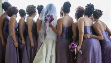 A bride stands looking out a window with her bridesmaids on both sides of her