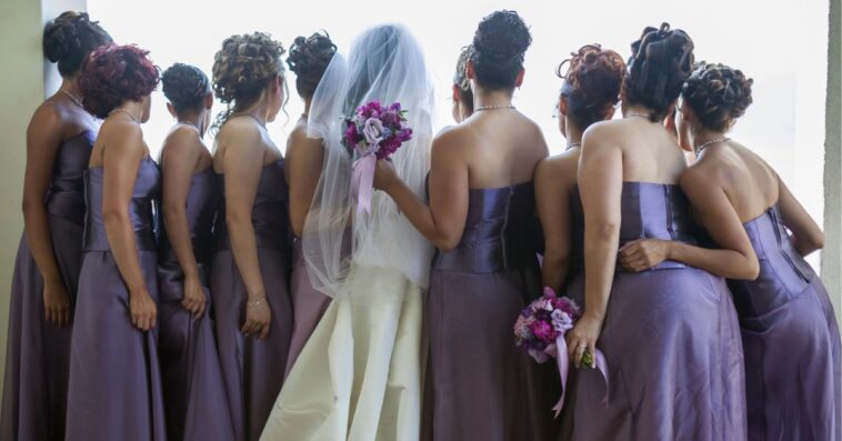 A bride stands looking out a window with her bridesmaids on both sides of her