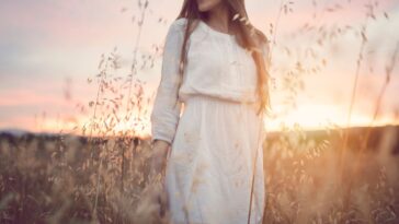 A young woman in a white dress stands in a field with the sun setting behind her
