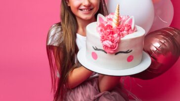 A young girl holds up a unicorn birthday cake, with pink balloons, and stands in front of a pink wall