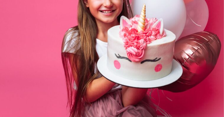 A young girl holds up a unicorn birthday cake, with pink balloons, and stands in front of a pink wall