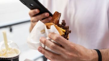 A man's hand holds a fast food sandwich with meat, bacon and cheese, the other hand holds an Iphone