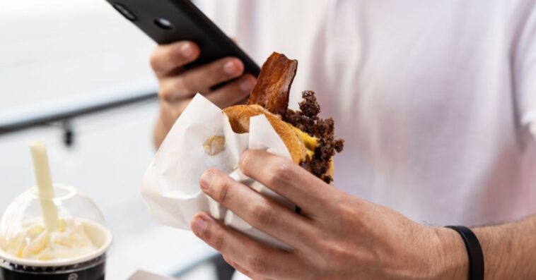 A man's hand holds a fast food sandwich with meat, bacon and cheese, the other hand holds an Iphone